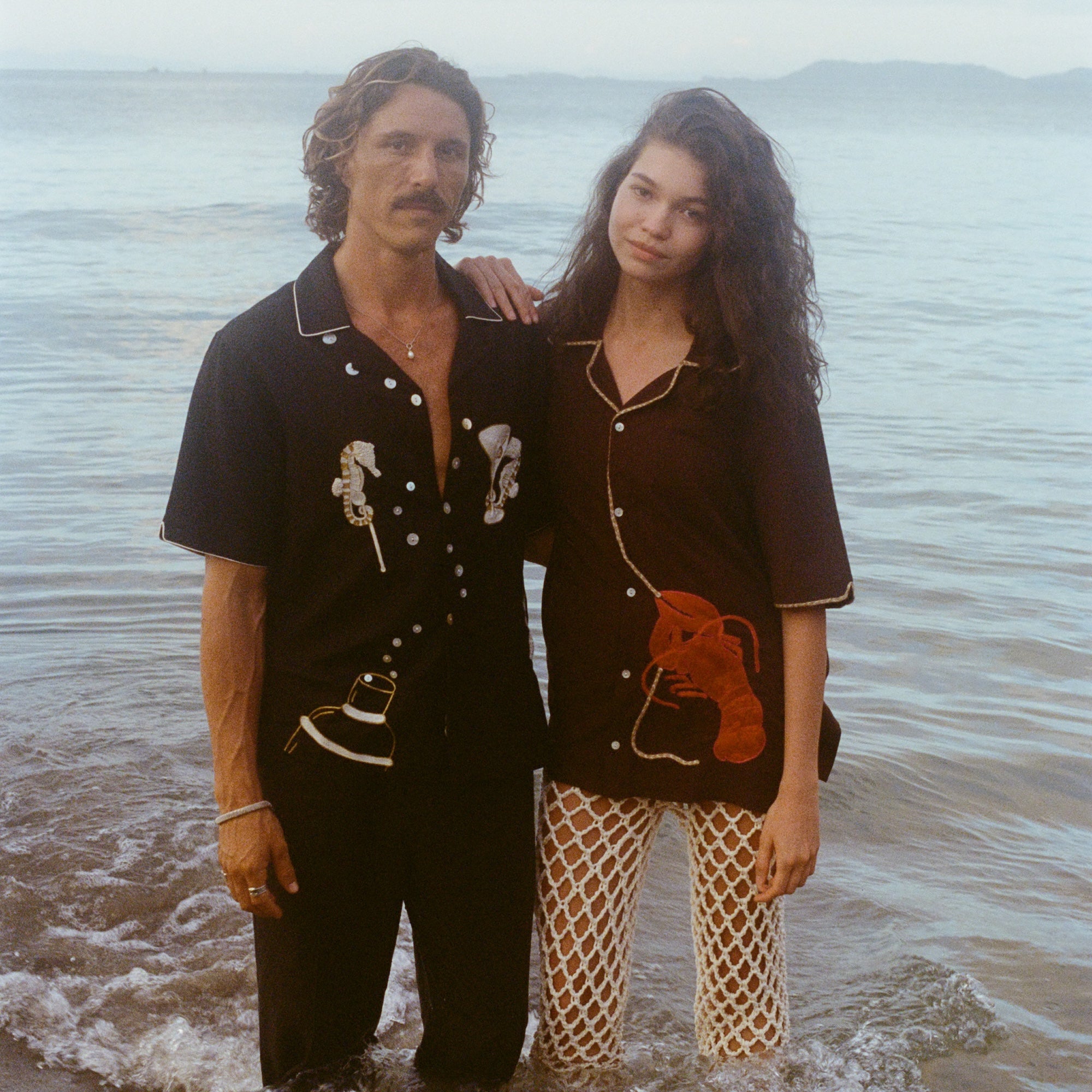 Two models stand ankle-deep in ocean water at sunset. The male model wears a black short-sleeve button-down shirt embroidered with two seahorses ('Tip your Bartenders' Cabana), paired with matching black trousers. The female model wears a brown short-sleeve shirt with beige piping and a large red lobster applique on the lower front ("Bisque Averse"), paired with white open-knit pants. They stand close together with the woman's hand resting gently on the man's shoulder.