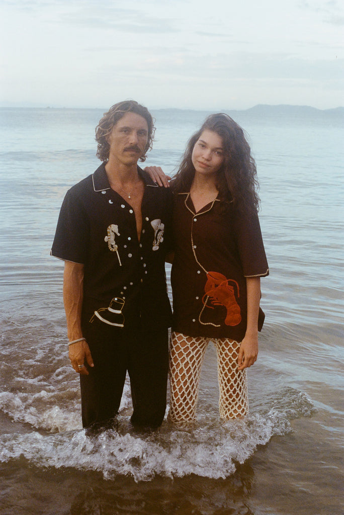 Two models stand ankle-deep in ocean water at sunset. The male model wears a black short-sleeve button-down shirt embroidered with two seahorses ('Tip your Bartenders' Cabana), paired with matching black trousers. The female model wears a brown short-sleeve shirt with beige piping and a large red lobster applique on the lower front ("Bisque Averse"), paired with white open-knit pants. They stand close together with the woman's hand resting gently on the man's shoulder.