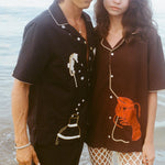 Cropped view of a man and woman standing close together at the shoreline, wearing short-sleeve button-up shirts with embroidery. The man’s black shirt features seahorses and a cocktail shaker; the woman’s brown shirt has a large red lobster design. Ocean water ripples in the background.