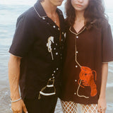 Cropped view of a man and woman standing close together at the shoreline, wearing short-sleeve button-up shirts with embroidery. The man’s black shirt features seahorses and a cocktail shaker; the woman’s brown shirt has a large red lobster design. Ocean water ripples in the background.