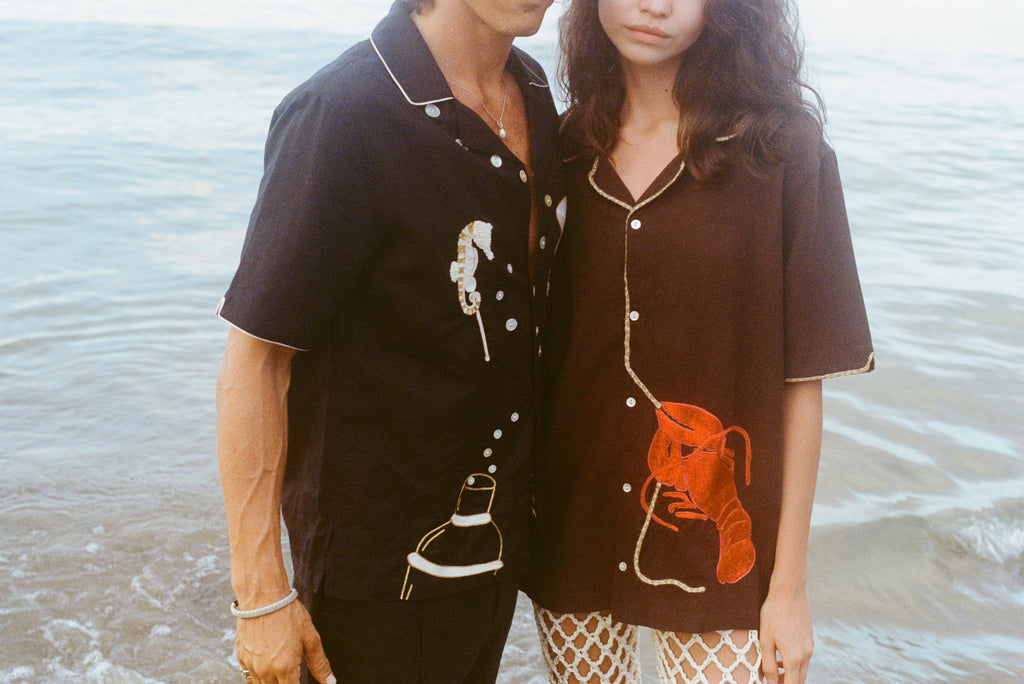 Cropped view of a man and woman standing close together at the shoreline, wearing short-sleeve button-up shirts with embroidery. The man’s black shirt features seahorses and a cocktail shaker; the woman’s brown shirt has a large red lobster design. Ocean water ripples in the background.