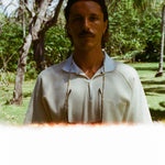 Man standing outdoors in a cream short-sleeve polo shirt with a white collar and V-notch chest pocket. Photo has a light leak effect at the bottom and trees in the background.