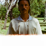 Man standing outdoors in a cream short-sleeve polo shirt with a white collar and V-notch chest pocket. Photo has a light leak effect at the bottom and trees in the background.