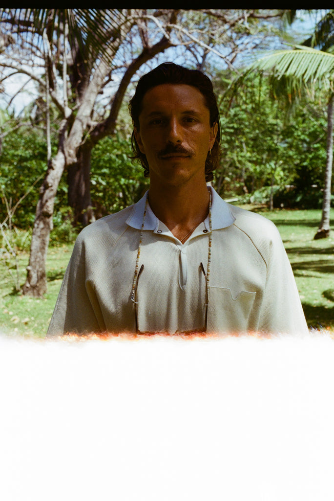 Man standing outdoors in a cream short-sleeve polo shirt with a white collar and V-notch chest pocket. Photo has a light leak effect at the bottom and trees in the background.