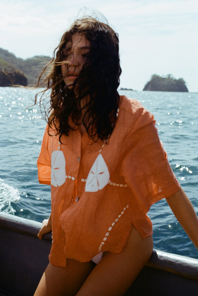 Image of brunette woman wearing the coral 'Sand Dollar Stimulus' ~ Relaxed Crop Cut, sitting on a boat and looking off to the side, with her hair blowing in the wind.