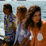 3 people sitting on a boat looking in different directions. Both women are wearing the 'Sand Dollar Stimulus' Shirt in both colorways (light blue and orange) while the man is wearing a patterned hawaiian shirt.