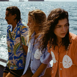 3 people sitting on a boat looking in different directions. Both women are wearing the 'Sand Dollar Stimulus' Shirt in both colorways (light blue and orange) while the man is wearing a patterned hawaiian shirt.