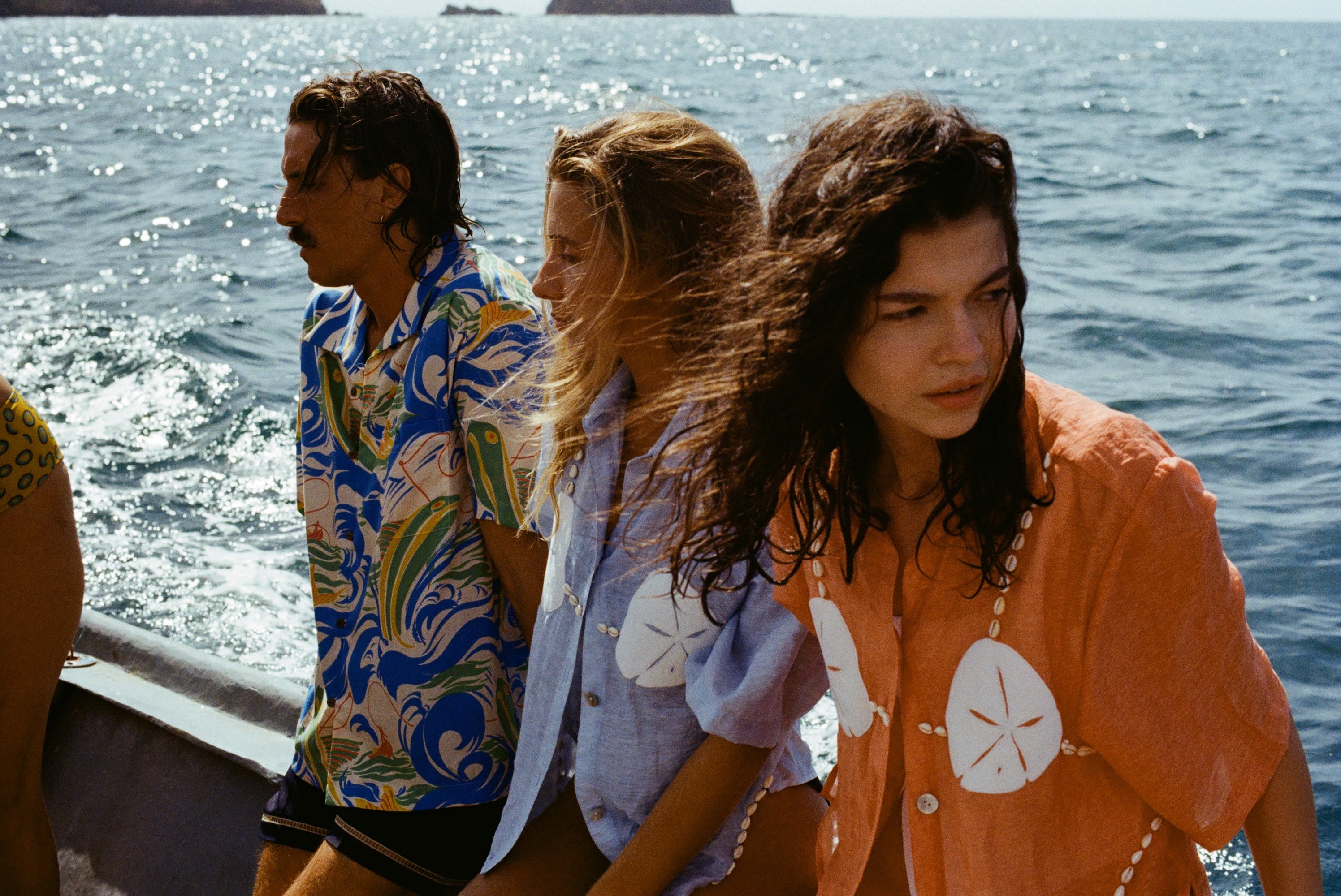 3 people sitting on a boat looking in different directions. Both women are wearing the 'Sand Dollar Stimulus' Shirt in both colorways (light blue and orange) while the man is wearing a patterned hawaiian shirt.