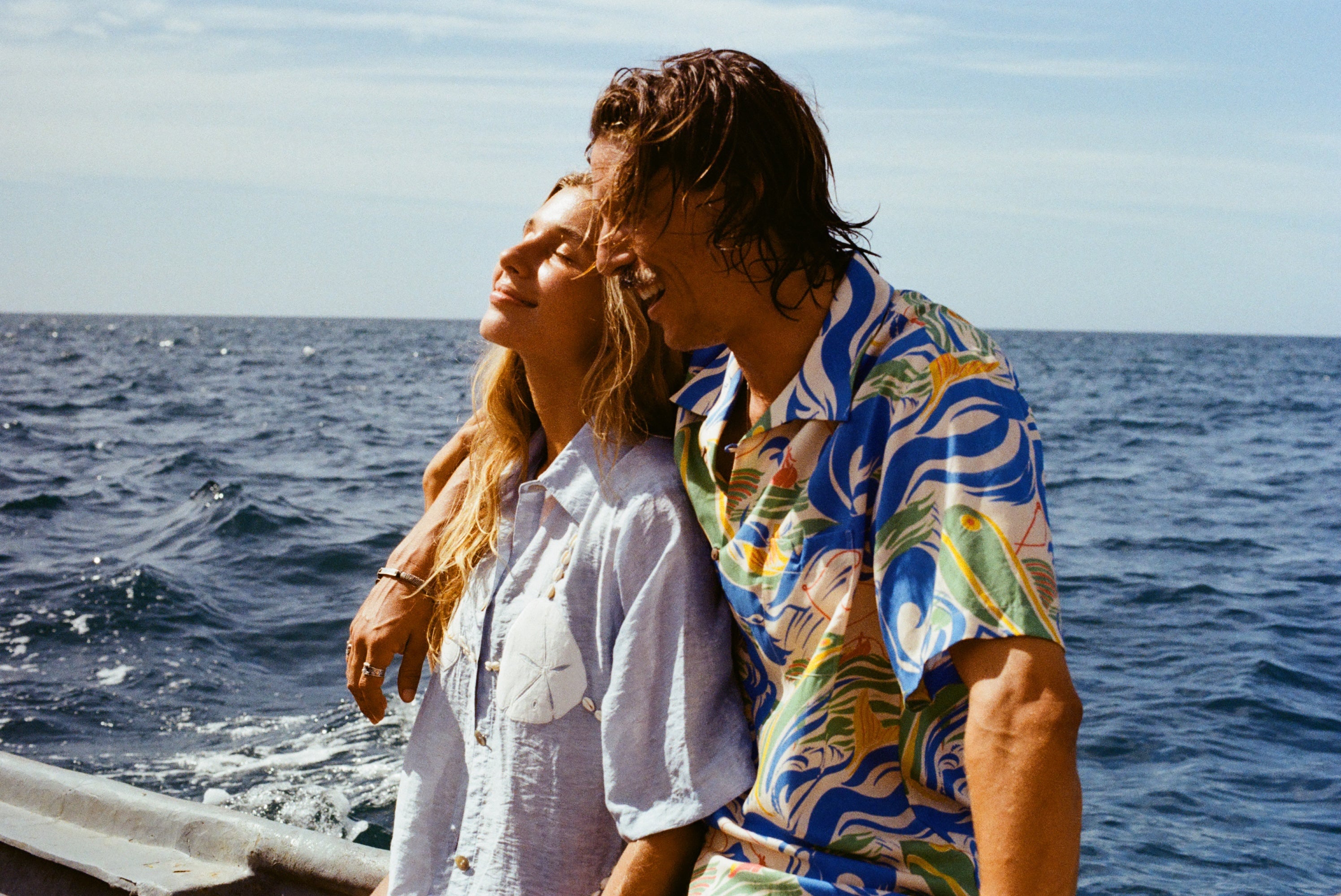 Male model wearing our 'Perfect Cast' Shirt, with his arm around a female model wearing the light blue sand dollar bikini shirt. Both are smiling while riding a boat.