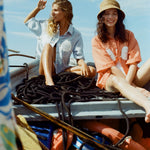 Two women sit on a boat surrounded by coiled black ropes and life vests. The woman on the left wears the light blue button-up bikini shirt, and is shading her eyes as she gazes into the distance. The woman on the right wears an orange version of the same shirt with a straw hat, smiling toward the camera. 