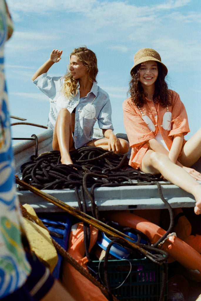 Two women sit on a boat surrounded by coiled black ropes and life vests. The woman on the left wears the light blue button-up bikini shirt, and is shading her eyes as she gazes into the distance. The woman on the right wears an orange version of the same shirt with a straw hat, smiling toward the camera. 