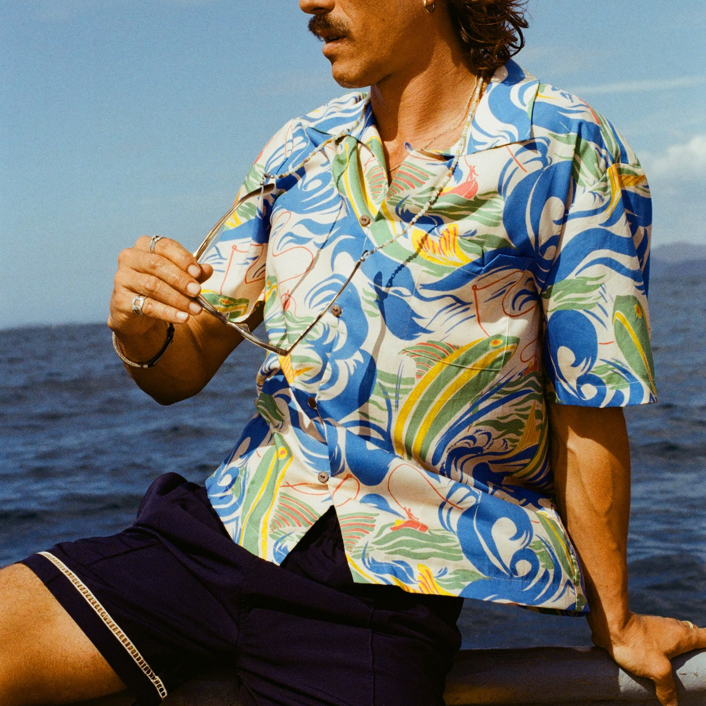 A man with wavy brown hair and a mustache sits on the edge of a boat, wearing a vibrant short-sleeve button-up shirt with a fishing-themed print featuring leaping fish and swirling ocean waves in blue, green, yellow, and red (Our 'Perfect Cast' Shirt). He pairs it with navy shorts that have white embroidered trim. He holds sunglasses in one hand and gazes toward the right.