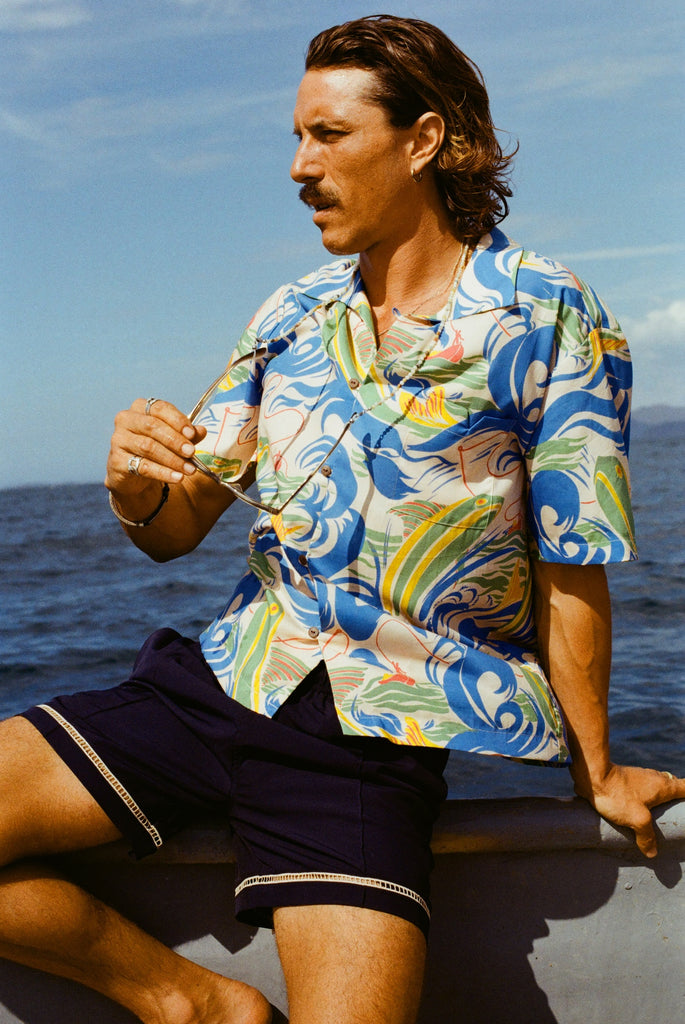 A man with wavy brown hair and a mustache sits on the edge of a boat, wearing a vibrant short-sleeve button-up shirt with a fishing-themed print featuring leaping fish and swirling ocean waves in blue, green, yellow, and red (Our 'Perfect Cast' Shirt). He pairs it with navy shorts that have white embroidered trim. He holds sunglasses in one hand and gazes toward the right.