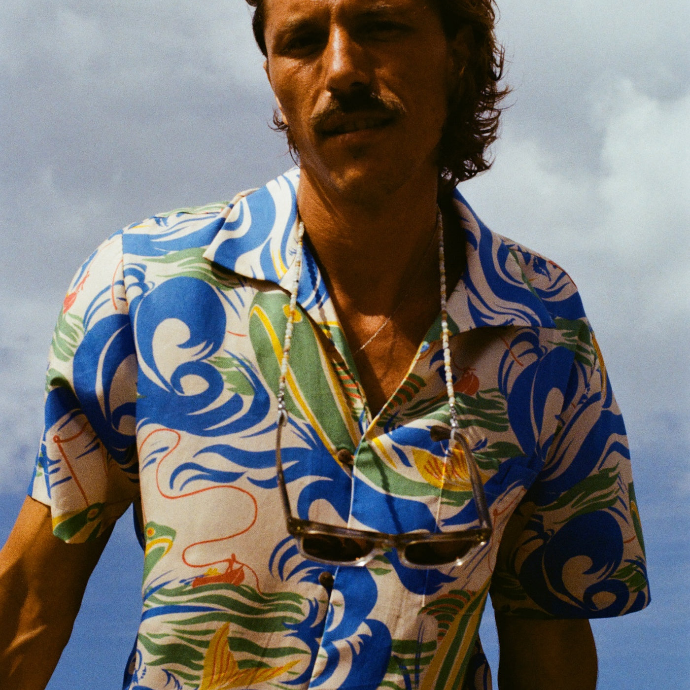 The same male model with longer hair and a mustache is staring into the camera, wearing our patterned 'Perfect Cast' Shirt and a pair of sunglasses are strapped around his neck. 