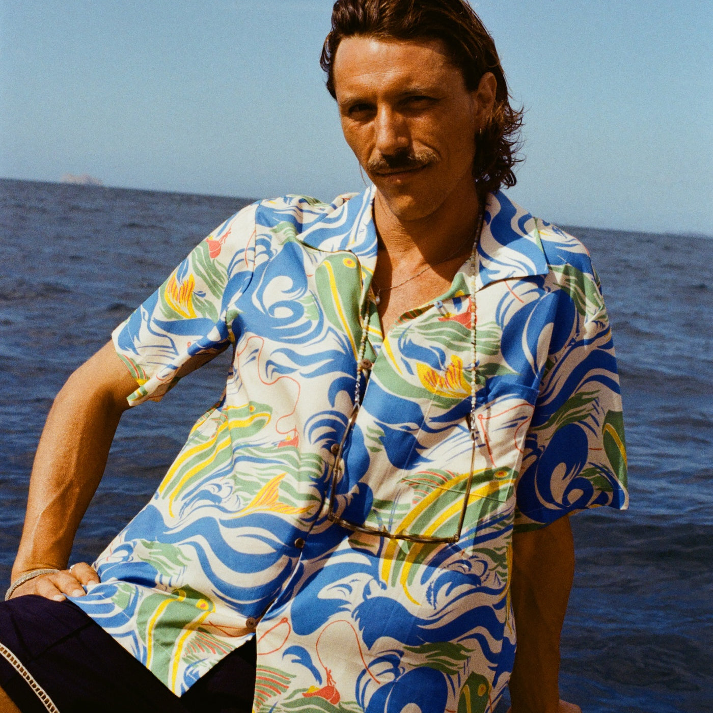 A man with wavy brown hair and a mustache sits on the edge of a boat, wearing a vibrant short-sleeve button-up shirt with a fishing-themed print featuring leaping fish and swirling ocean waves in blue, green, yellow, and red (Our 'Perfect Cast' Shirt). He pairs it with navy shorts that have white embroidered trim. 