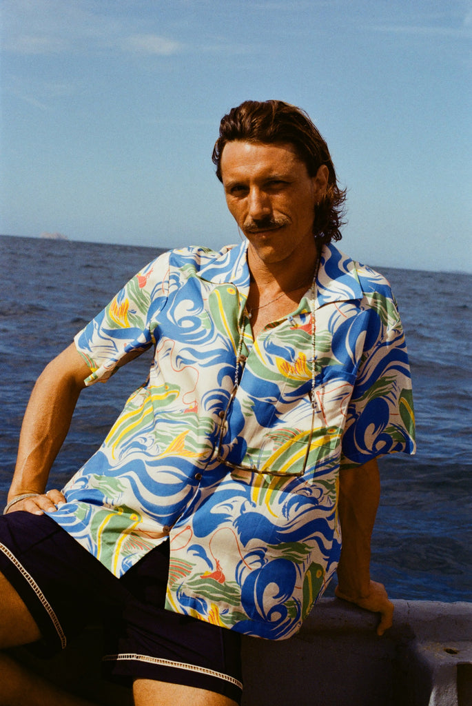 A man with wavy brown hair and a mustache sits on the edge of a boat, wearing a vibrant short-sleeve button-up shirt with a fishing-themed print featuring leaping fish and swirling ocean waves in blue, green, yellow, and red (Our 'Perfect Cast' Shirt). He pairs it with navy shorts that have white embroidered trim. 