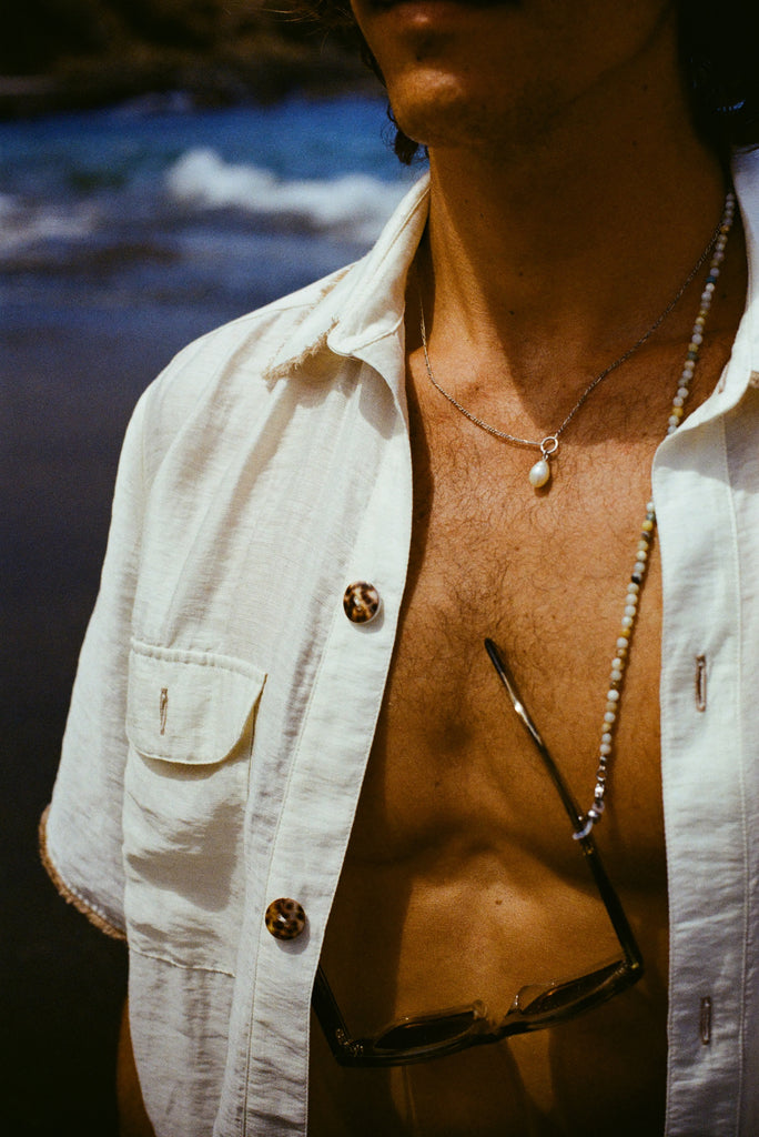 Close up of a man's chest, wearing a white shirt open, a necklace with a pearl and sunglasses with a beaded sunglasses chain