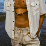 Man wearing a button down shirt open, with sunglasses hanging, beige shorts, and hands in pockets.