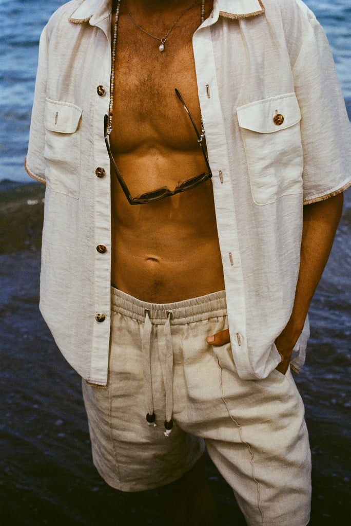 Man wearing a button down shirt open, with sunglasses hanging, beige shorts, and hands in pockets.