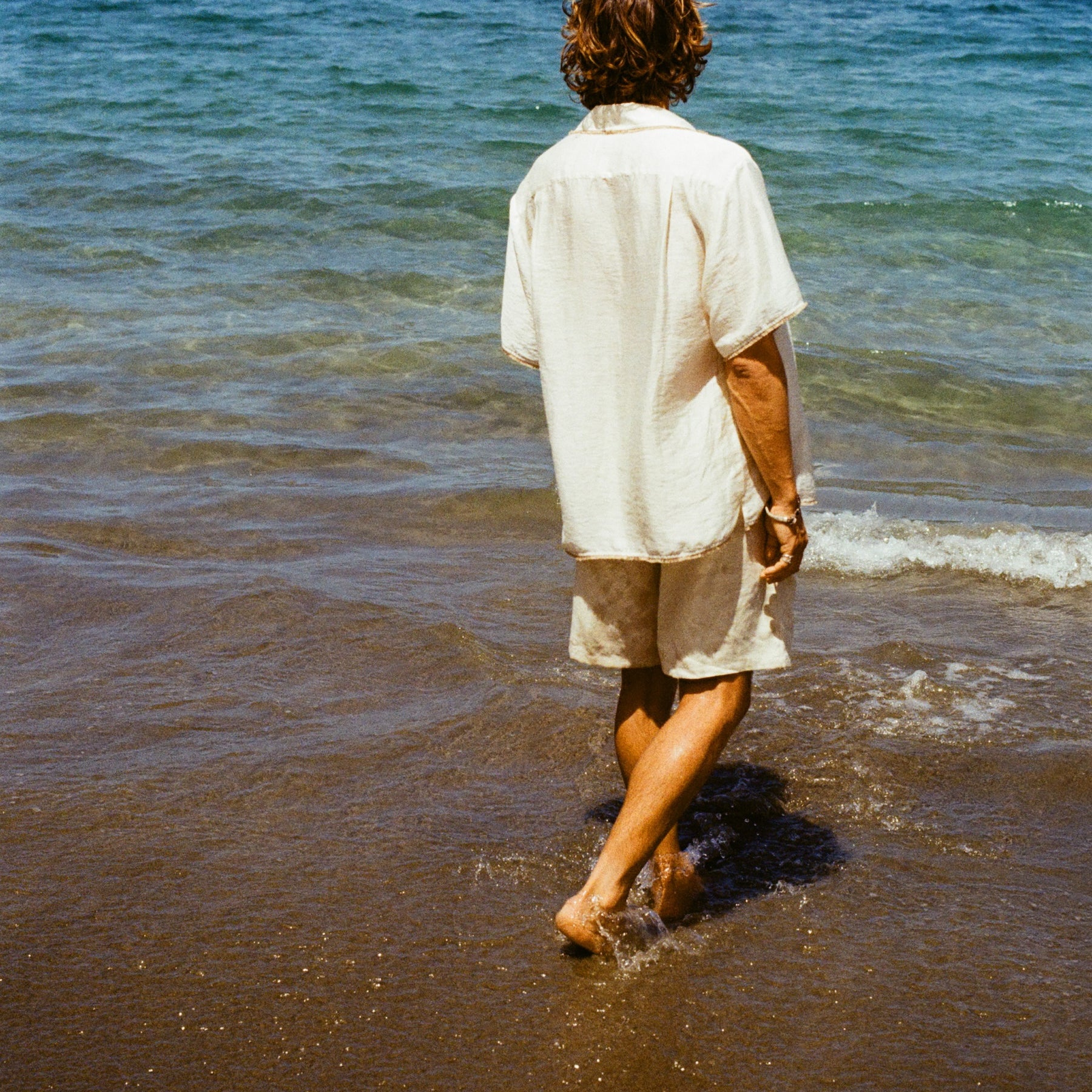 Man at the beach facing away from the camera walking towards the ocean, wearing a white shirt and beige shorts