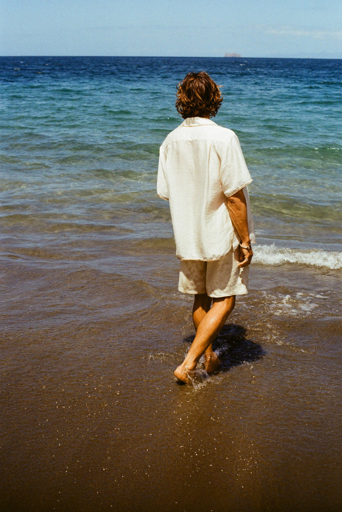 Man at the beach facing away from the camera walking towards the ocean, wearing a white shirt and beige shorts