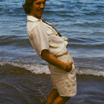 Brunette man standing in the ocean, wearing the sand linen Easy Going Shorts and our featherweight Resort Shirt and leaning backwards, with his hands in his pocket. 