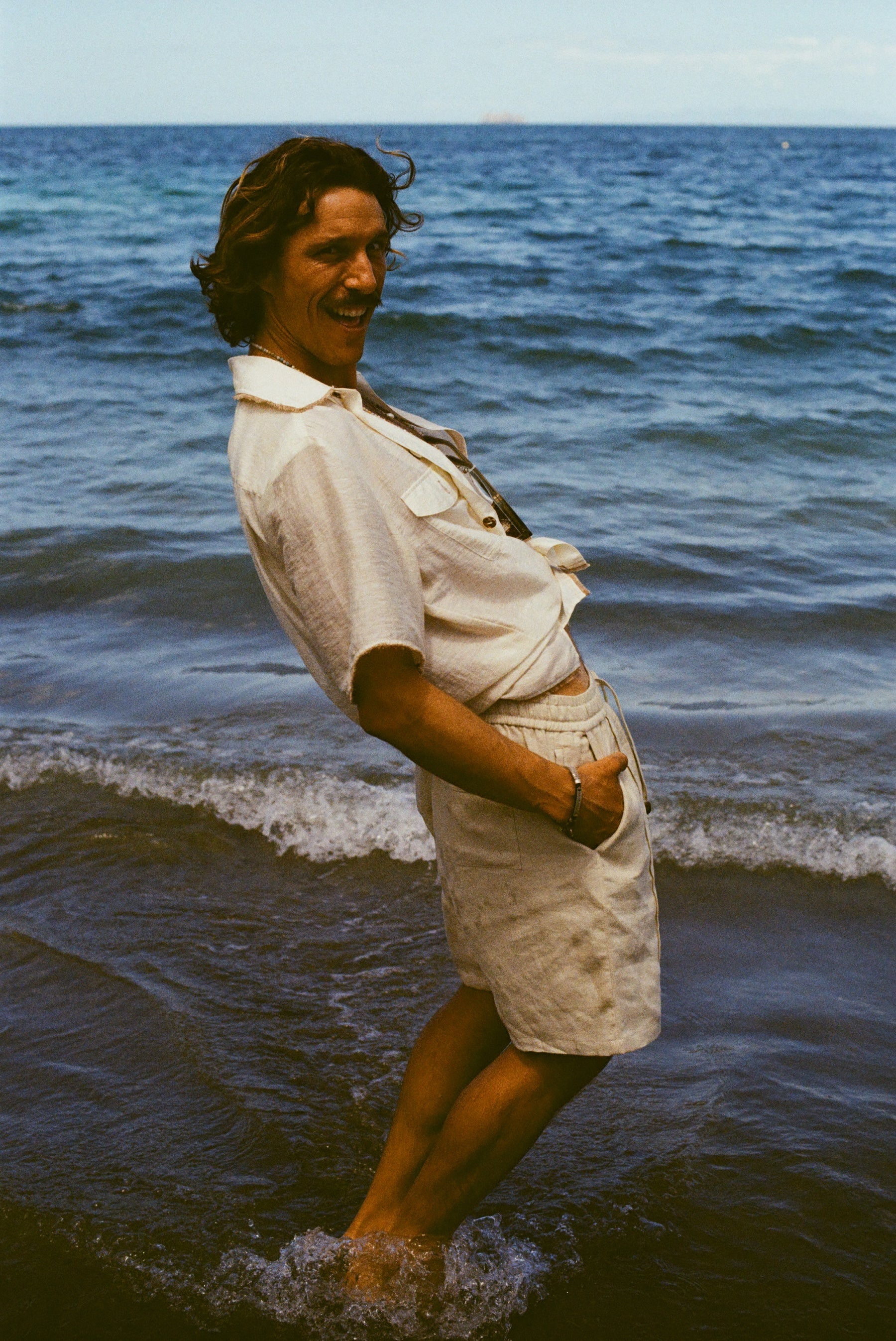 Man at the beach, facing the camera and leaning back, smiling, wearing a white button down shirt and beige linen shorts