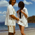 Image of man and woman standing on sandy beach, both wearing the 'Seasons of Change' by Mary Ball Shirts, in green and blue. 