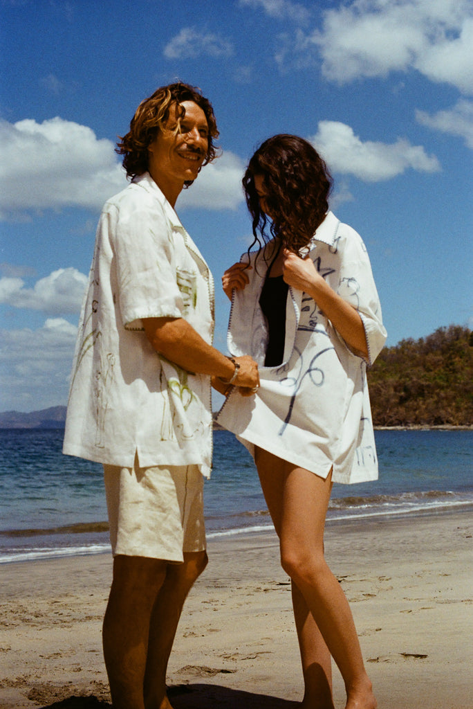 Image of man and woman standing on sandy beach, both wearing the 'Seasons of Change' by Mary Ball Shirts, in green and blue. 