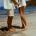 Image of man and woman standing on sandy beach, both wearing the 'Seasons of Change' by Mary Ball Shirts, in green and blue. 