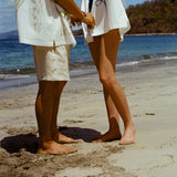 Image of man and woman standing on sandy beach, both wearing the 'Seasons of Change' by Mary Ball Shirts, in green and blue. 