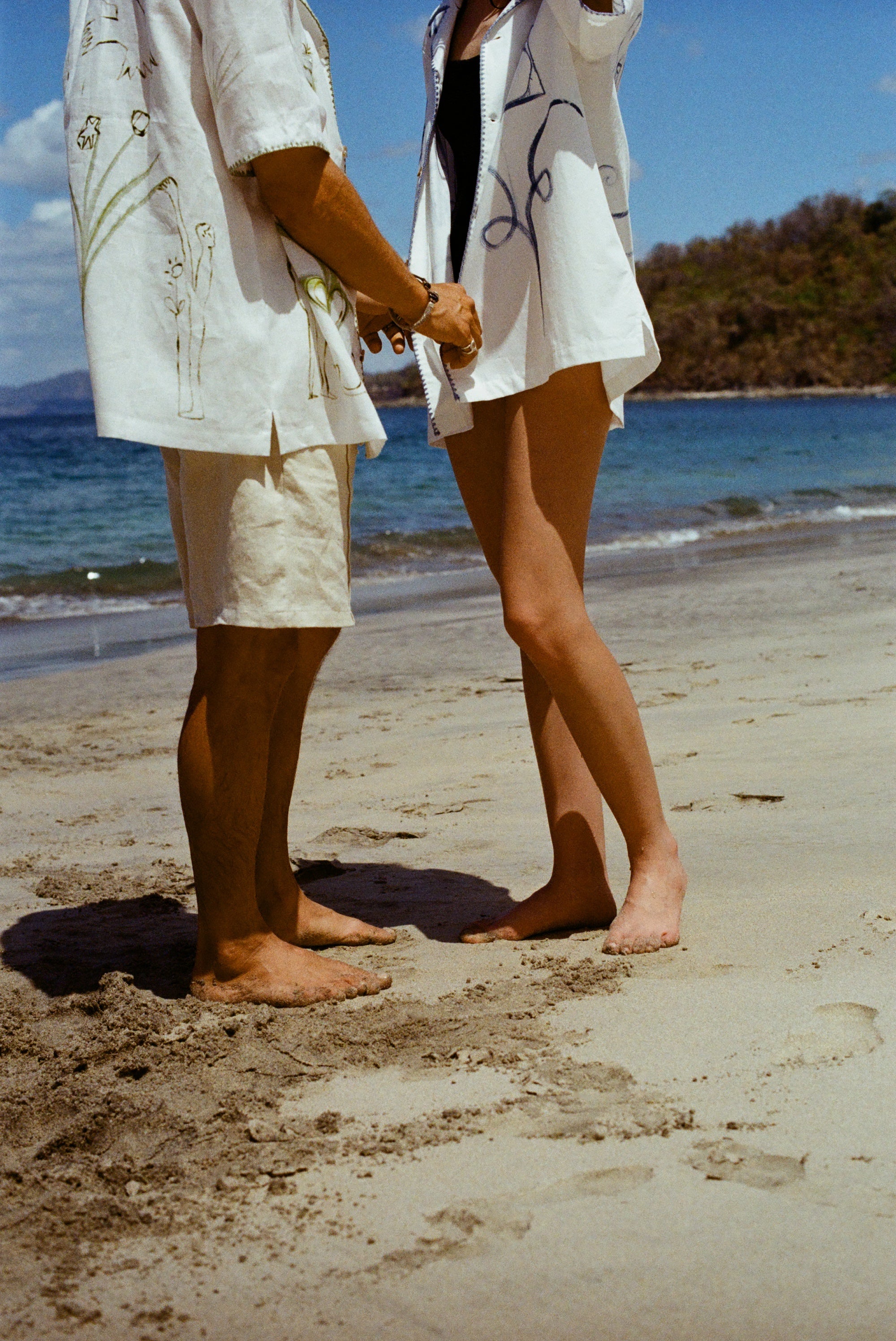 Image of man and woman standing on sandy beach, both wearing the 'Seasons of Change' by Mary Ball Shirts, in green and blue. 