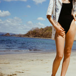 Cropped image of woman in black swimsuit and the 'Seasons of Change' by Mary Ball Shirt unbuttoned on top, with her hand on hip.