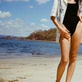 Cropped image of woman in black swimsuit and the 'Seasons of Change' by Mary Ball Shirt unbuttoned on top, with her hand on hip.