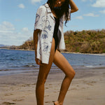 Image from the side of woman standing on beach in a black swimsuit, with the 'Seasons of Change' by Mary Ball Shirt unbuttoned over it. She has one heel popped and her am behind her head. 