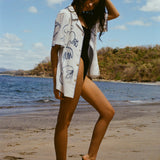 Image from the side of woman standing on beach in a black swimsuit, with the 'Seasons of Change' by Mary Ball Shirt unbuttoned over it. She has one heel popped and her am behind her head. 