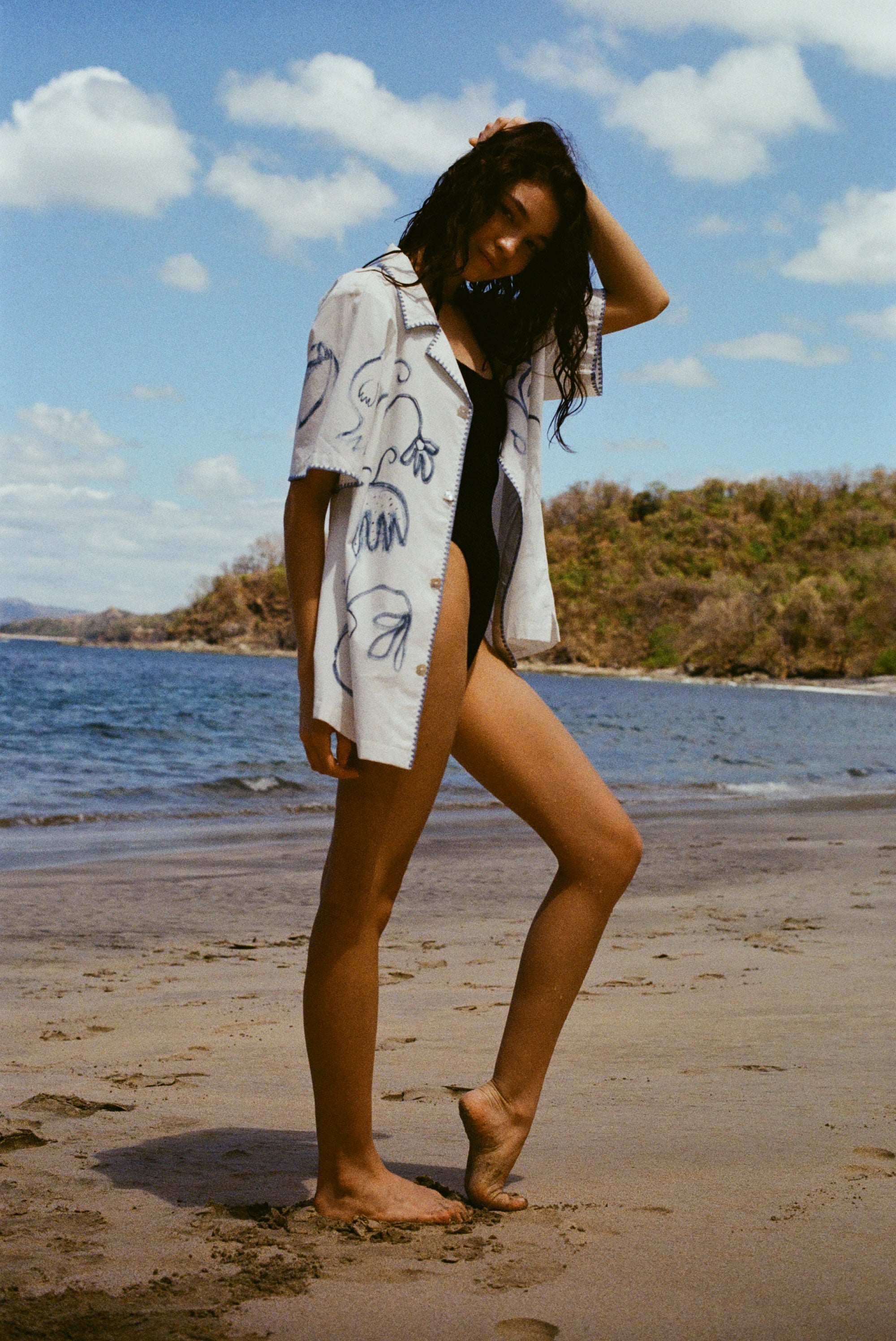 Image from the side of woman standing on beach in a black swimsuit, with the 'Seasons of Change' by Mary Ball Shirt unbuttoned over it. She has one heel popped and her am behind her head. 
