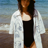 Woman on beach in black one piece bathing suit, wearing the 'Seasons of Change' by Mary Ball Shirt in Blue unbuttoned. 
