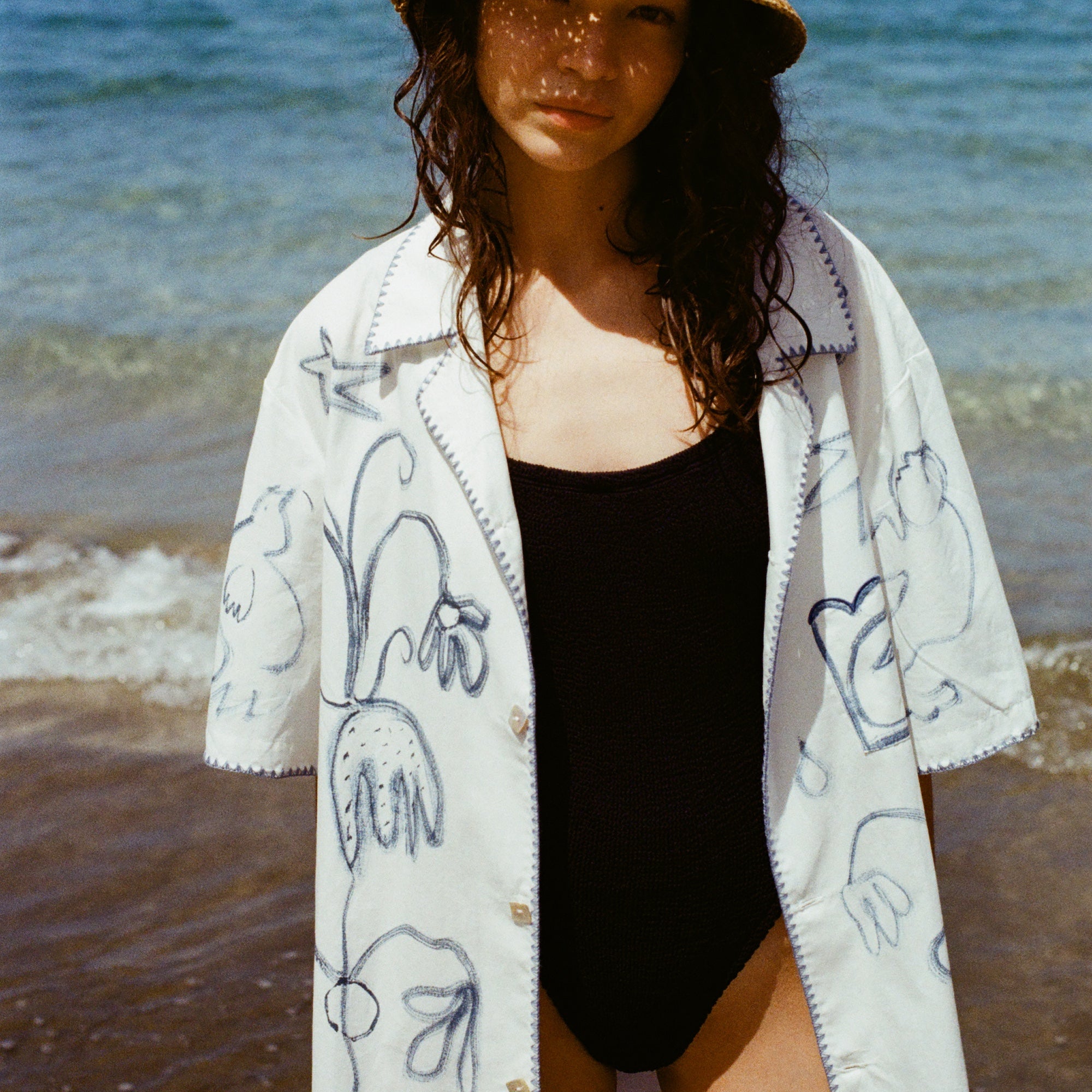 Woman on beach in black one piece bathing suit, wearing the 'Seasons of Change' by Mary Ball Shirt in Blue unbuttoned. 