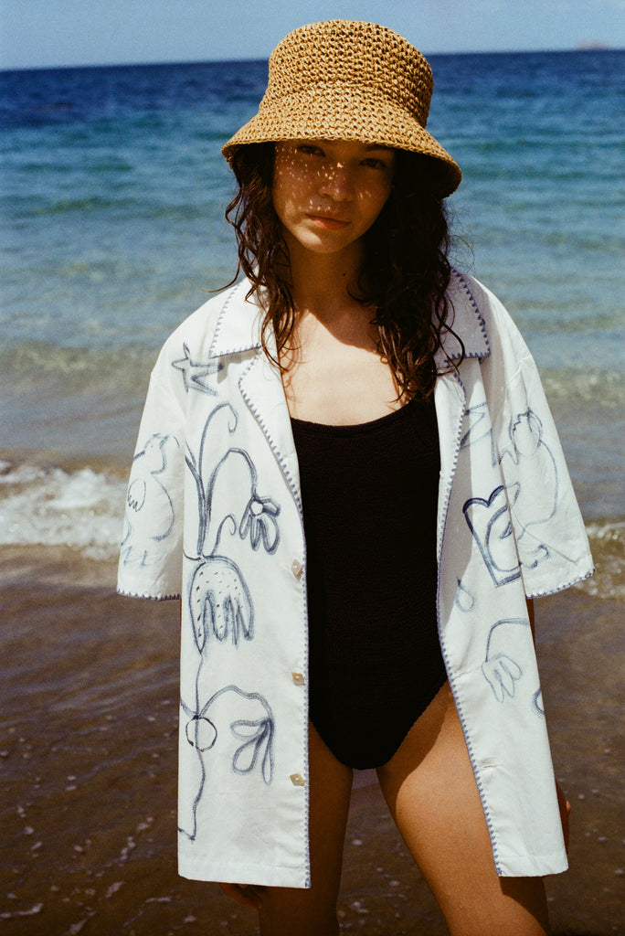 Woman on beach in black one piece bathing suit, wearing the 'Seasons of Change' by Mary Ball Shirt in Blue unbuttoned. 
