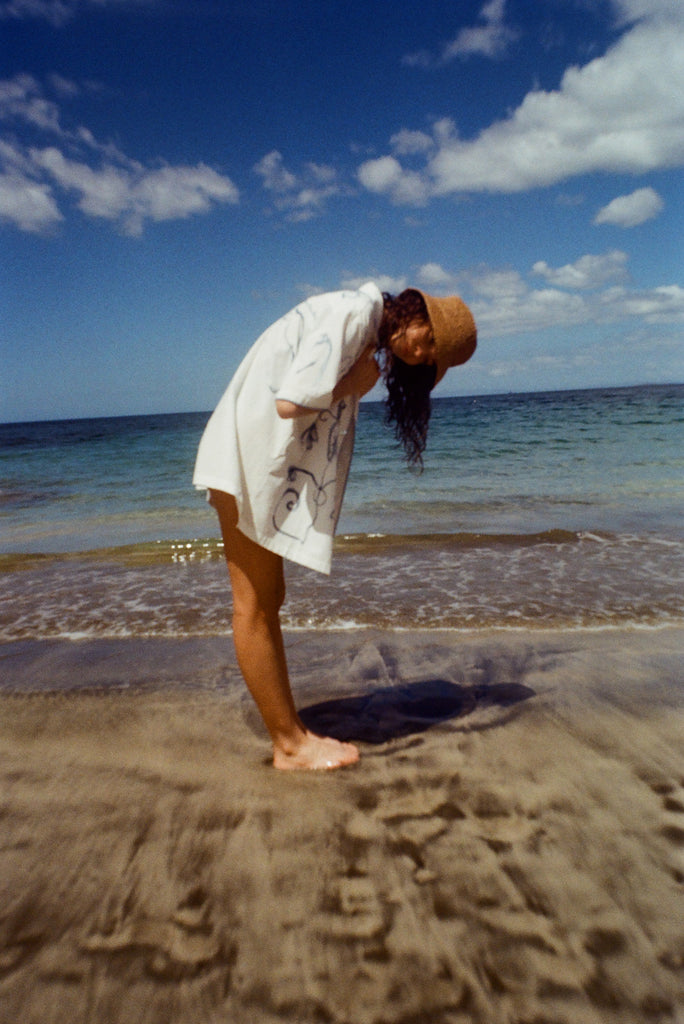 Woman on beach standing with her side to us, wearing the 'Seasons of Change' by Mary Ball Shirt and looking down into the sand. 