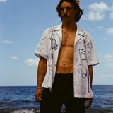 Man standing on beach wearing the 'Seasons of Change' by Mary Ball Shirt in blue unbuttoned, with our navy linen Easy Going Shorts. 