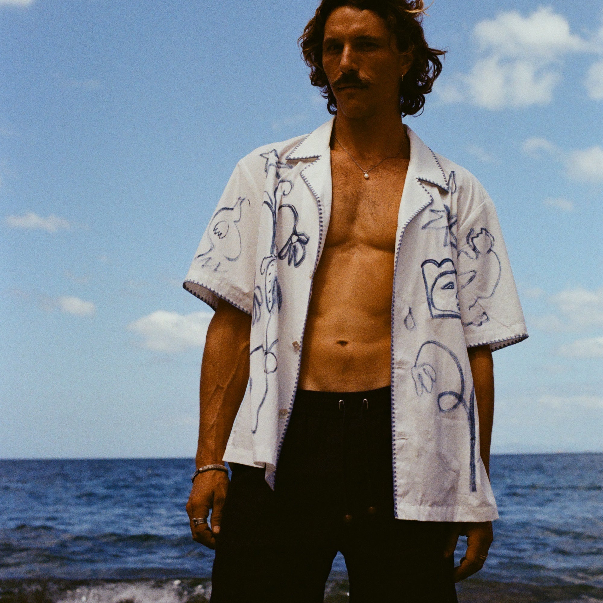 Man standing on beach wearing the 'Seasons of Change' by Mary Ball Shirt in blue unbuttoned, with our navy linen Easy Going Shorts. 
