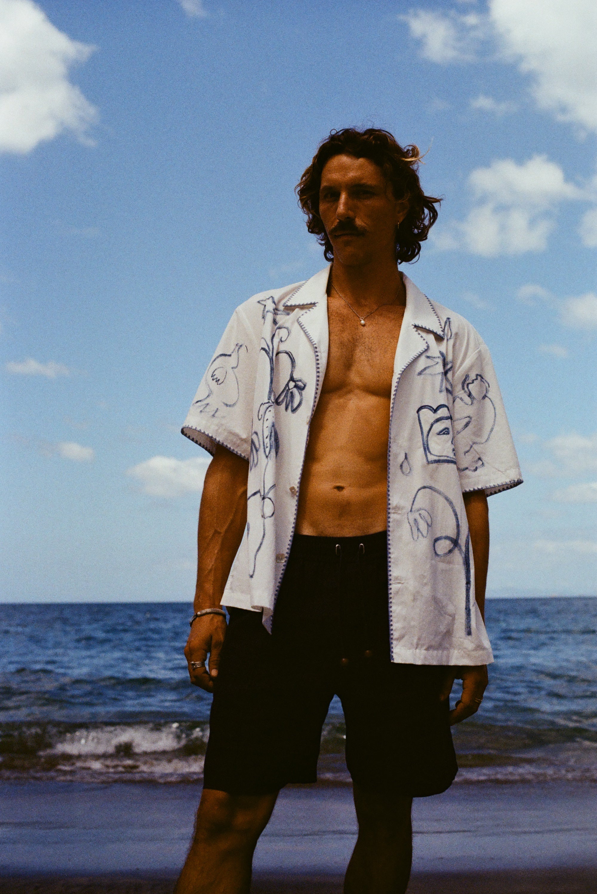 Man standing on beach wearing the 'Seasons of Change' by Mary Ball Shirt in blue unbuttoned, with our navy linen Easy Going Shorts. 