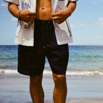 Close-up image of man standing on beach wearing the Navy Linen Easy Going Shorts, and our artist residency shirt unbuttoned. 
