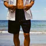 Close-up image of man standing on beach wearing the Navy Linen Easy Going Shorts, and our artist residency shirt unbuttoned. 