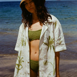 Woman standing on beach in green bikini, straw hat, and wearing the 'Seasons of Change' by Mary Ball Shirt in Green.
