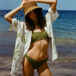 A woman standing on a beach wearing a green bikini, an open white shirt with green designs, and a brown sun hat.