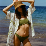 A woman standing on a beach wearing a green bikini, an open white shirt with green designs, and a brown sun hat.