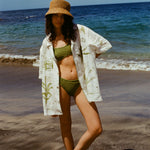 Woman standing on beach in green bikini, straw hat, and wearing the 'Seasons of Change' by Mary Ball Shirt in Green, with her hand on her hip.